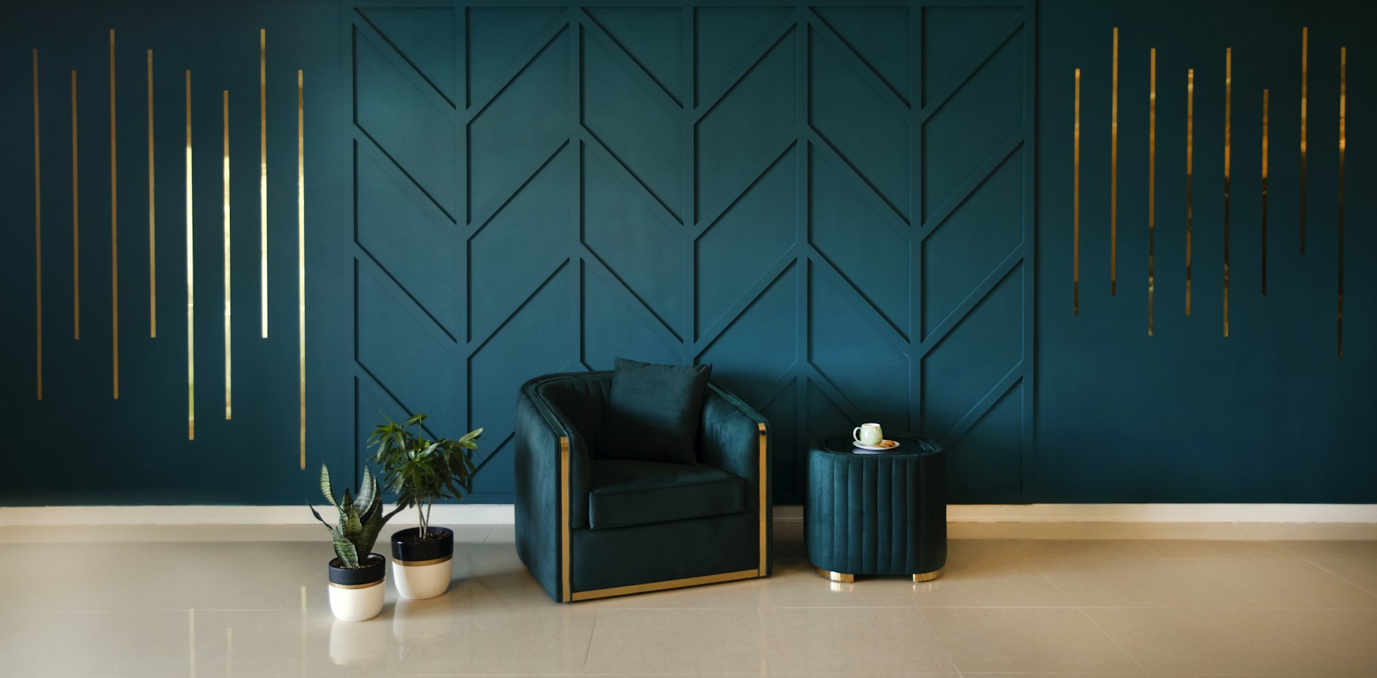 Teal wall with raised geometric panels and gold accents, a teal armchair, a small side table and two potted plants